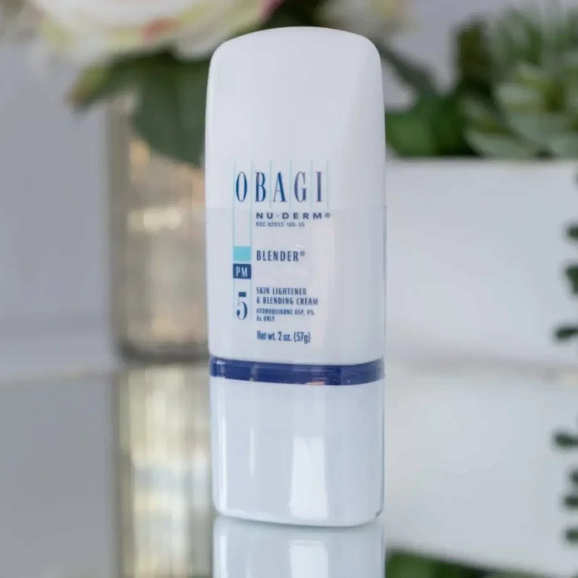 Obagi Nu-Derm Blender skin lightener cream bottle on reflective surface with plants in background