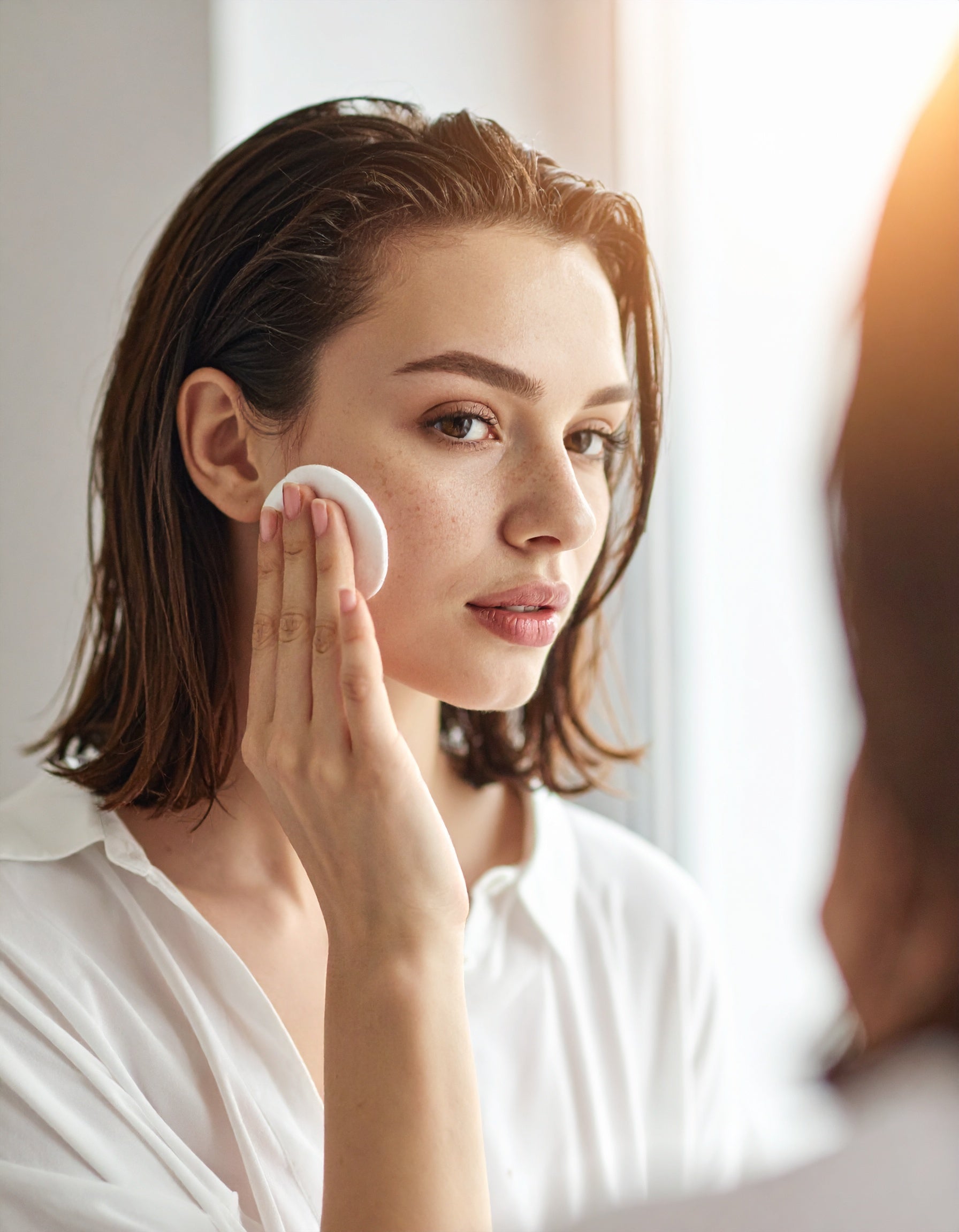Woman applying skincare with cotton pad, natural glow, freckled skin, morning routine
